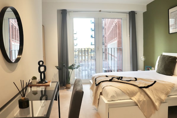 A modern hotel room with a large bed, a glass table, a mirror, a potted plant, and a view of the city through the balcony doors.