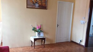 Een woonkamer met een houten vloer, een rode bank, een kleine tafel met een bloevas, een schilderij aan de muur en een deur.