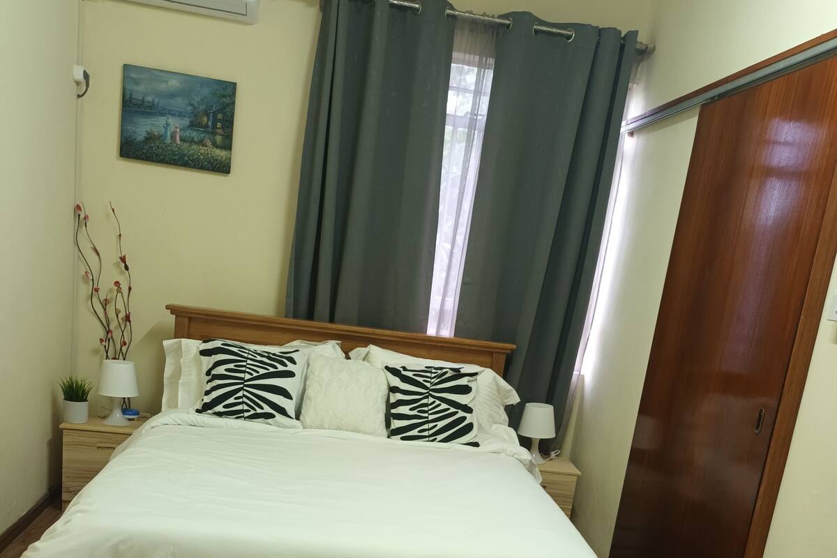 Een netjes opgemaakt bed met wit beddengoed en gestreepte kussens, een zacht wit vloerkleed, een houten hoofdbord, een nachtkastje met een lamp, een wandairco en een deur aan de rechterkant.
