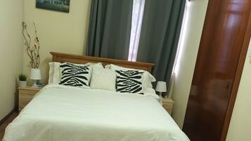 Una cama bien hecha con ropa blanca y almohadas estampadas, una alfombra blanca y esponjosa, un cabecero de madera, una mesita de noche con una lámpara, un aire acondicionado de pared y una puerta a la derecha.