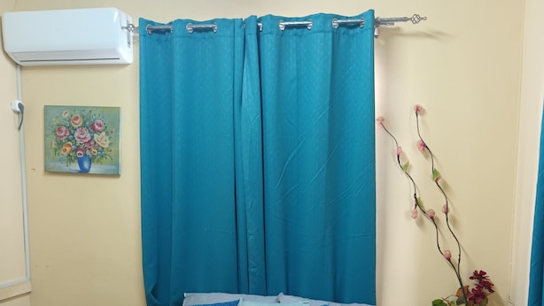 Een net opgemaakt bed met blauw-grijze beddengoed, een blauw gordijn, een klein nachtkastje met een lamp en een wandairconditioner.