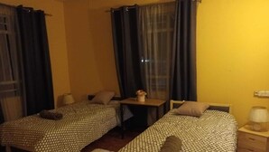 Een kamer met twee bedden, elk voorzien van een bedovertrek met dessin en kussens, een klein tafeltje met een lamp, en een raam met gordijnen.