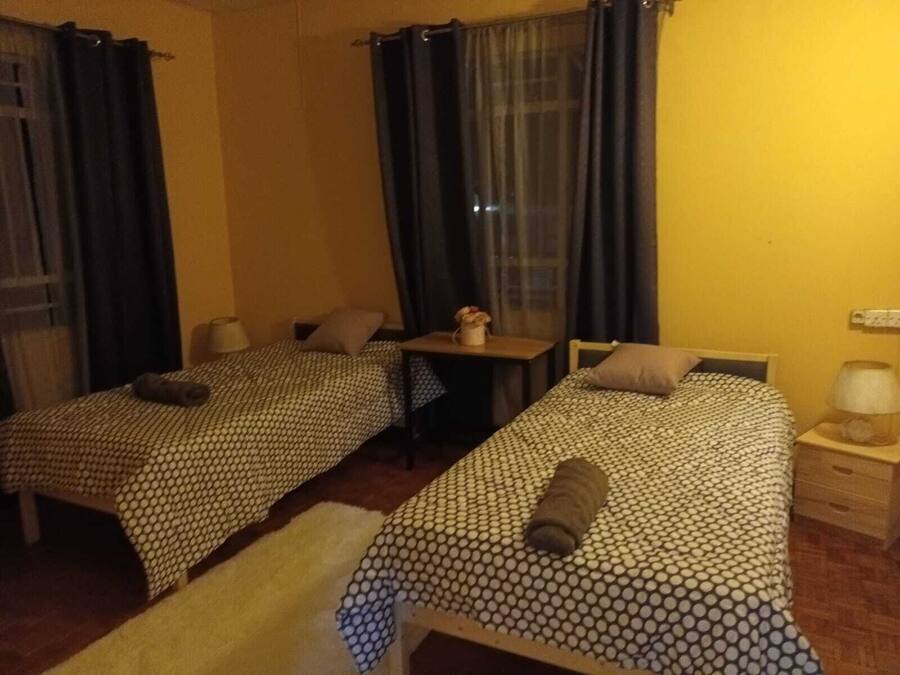 Ein Zimmer mit zwei Betten, jeweils mit gemustertem Überwurf und Kissen, einem kleinen Tisch mit Lampe sowie einem Fenster mit Vorhängen.