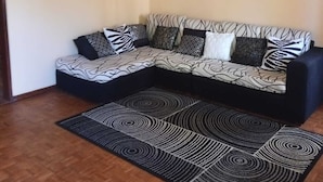 Een woonkamer met een hoekbank, gekleurde kussens, een zwart-wit kleed en wanddecoratie.