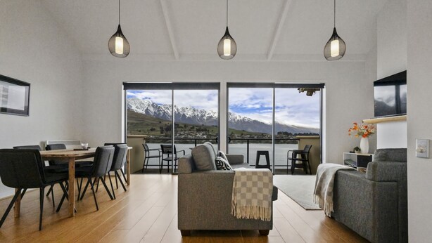 A modern living room with a large window overlooking a mountainous landscape, a dining area with a wooden table and chairs, and a cozy seating area with a sofa and armchairs. - Lakeside Landing - lakefront and walk to town (Queenstown)