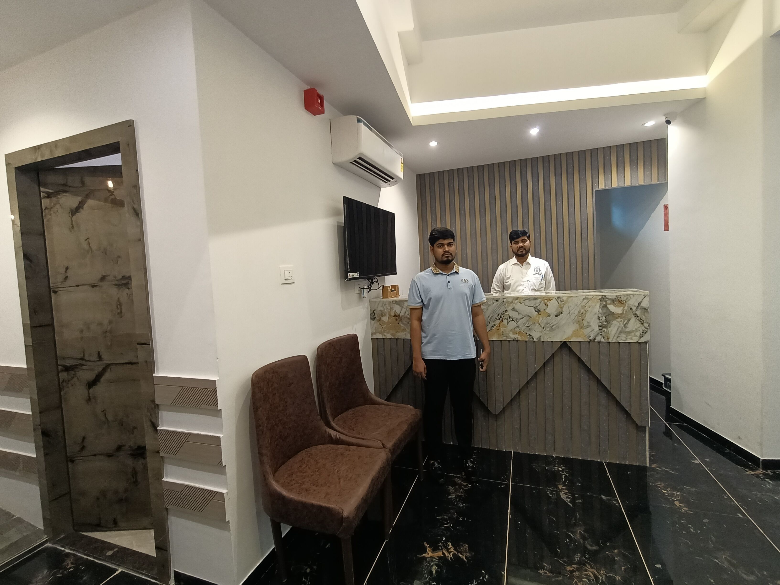 A reception area with a marble counter, two brown chairs, a mounted TV, and a red fire alarm.