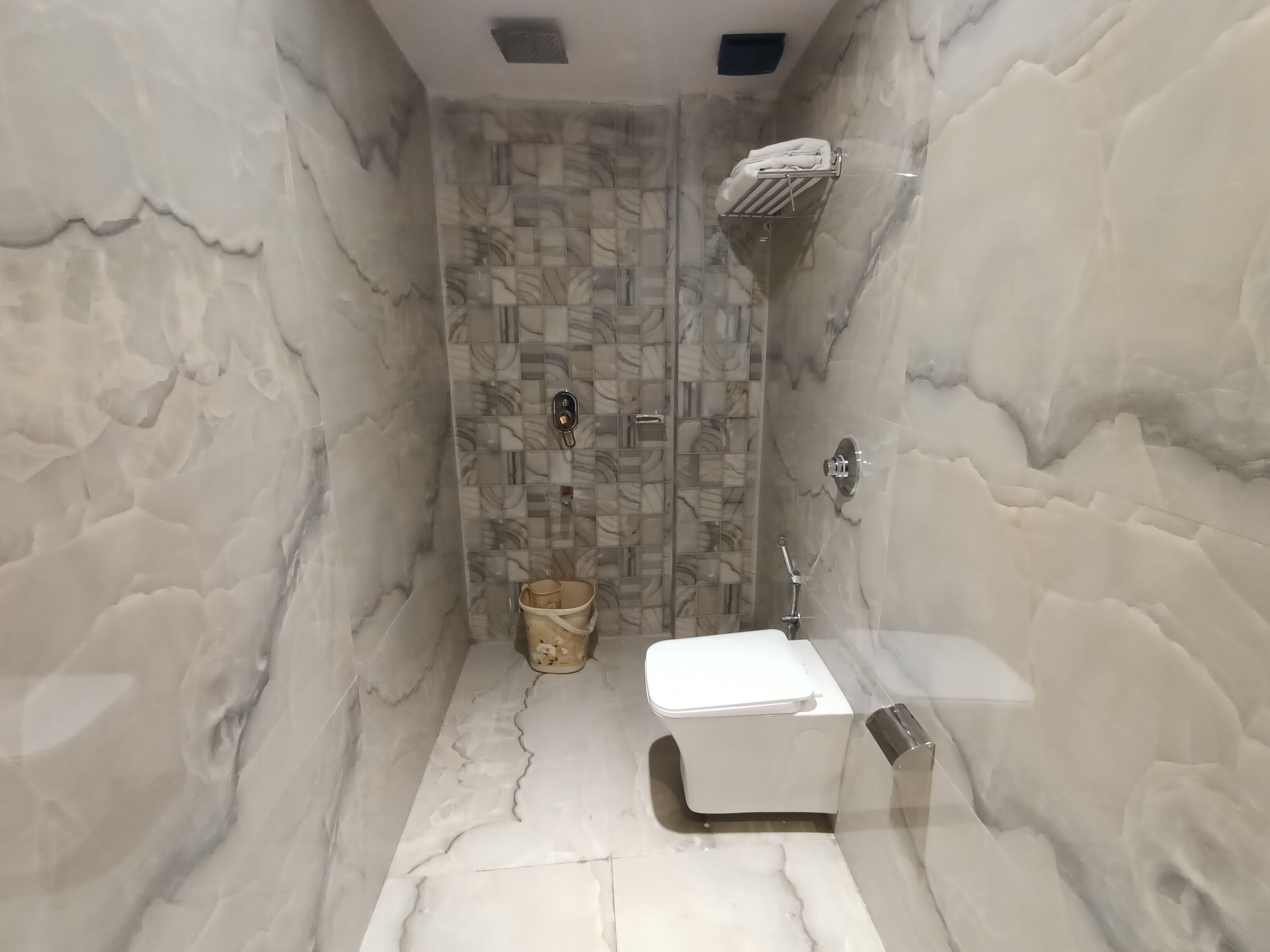 A bathroom with a marble wall, a shower, a toilet, and a small bucket.