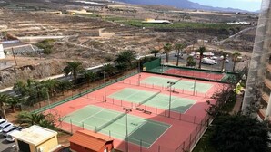 A tennis court complex with multiple courts, surrounded by a fence, and adjacent to a building with a red roof. - Marazul DEL SUR Face A LA MER (ADEJE)