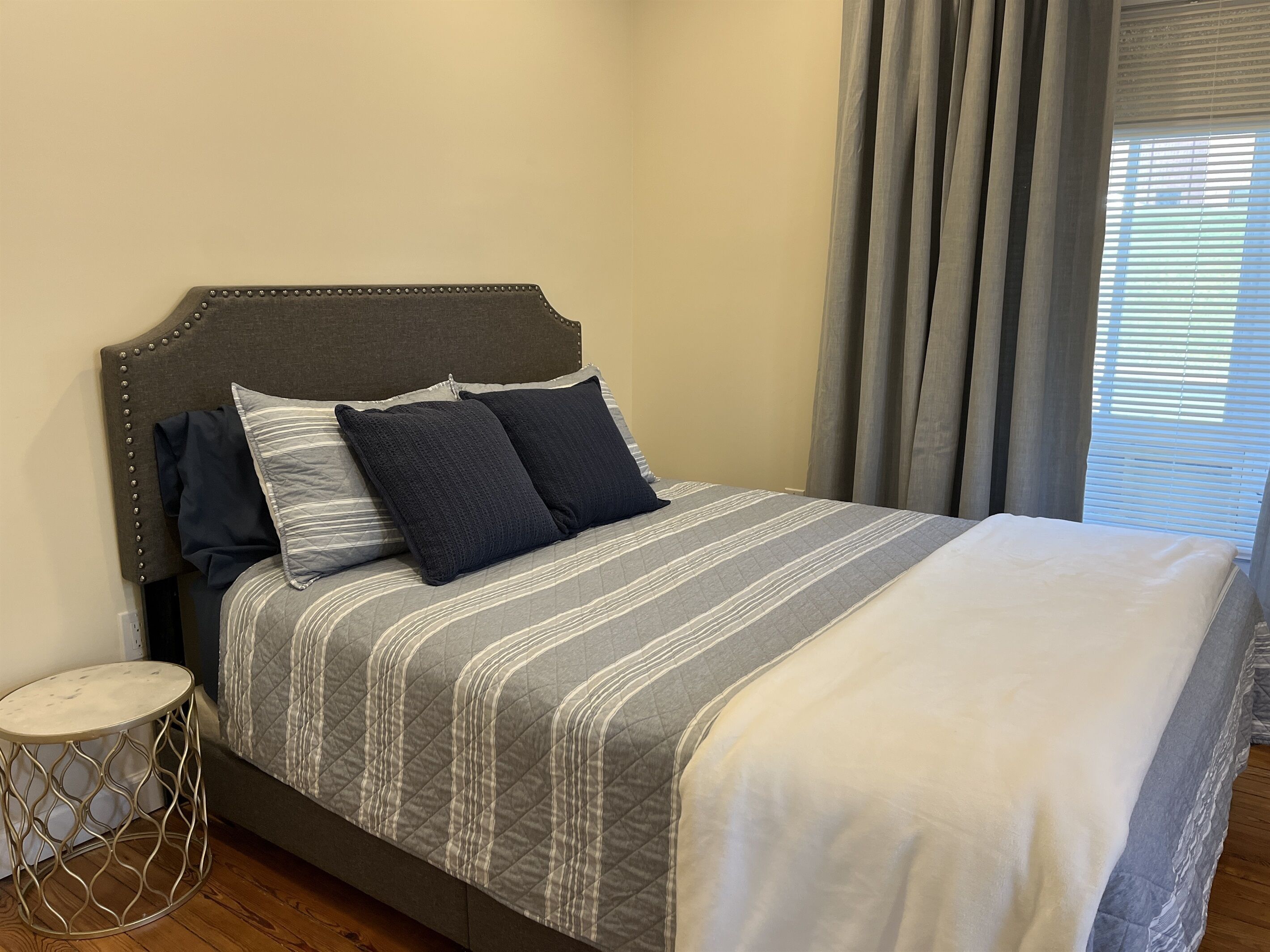 A neatly made bed with a striped bedspread and two pillows, a round metal side table, and a window with curtains.