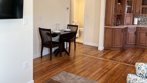 A dining area with a wooden table and chairs, a TV mounted on the wall, and a kitchen with wooden cabinets and a tiled backsplash. - BigWave 1 -Total reno 1st fl, near ASV & Colleges (Oneonta)