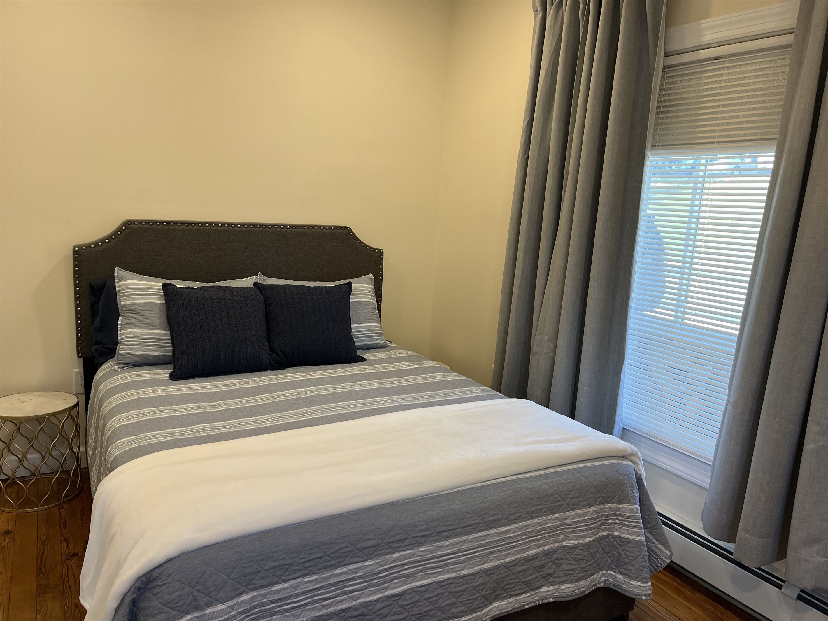 A neatly made bed with a striped bedspread and pillows, a wooden nightstand with a decorative item, and a window with blinds.