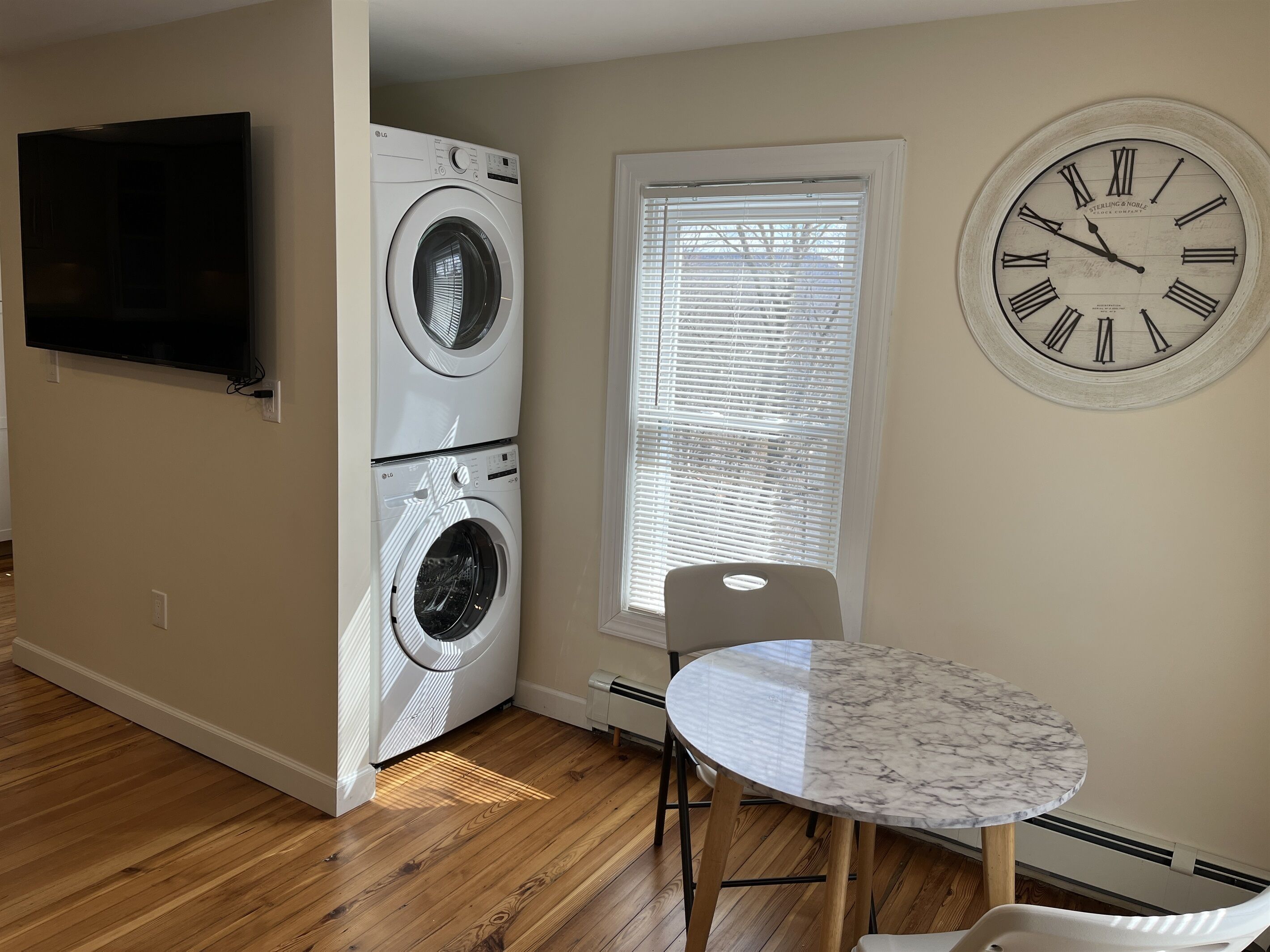 A laundry room with two stacked washing machines, a television mounted on the wall, a window with blinds, and a round table with two chairs.