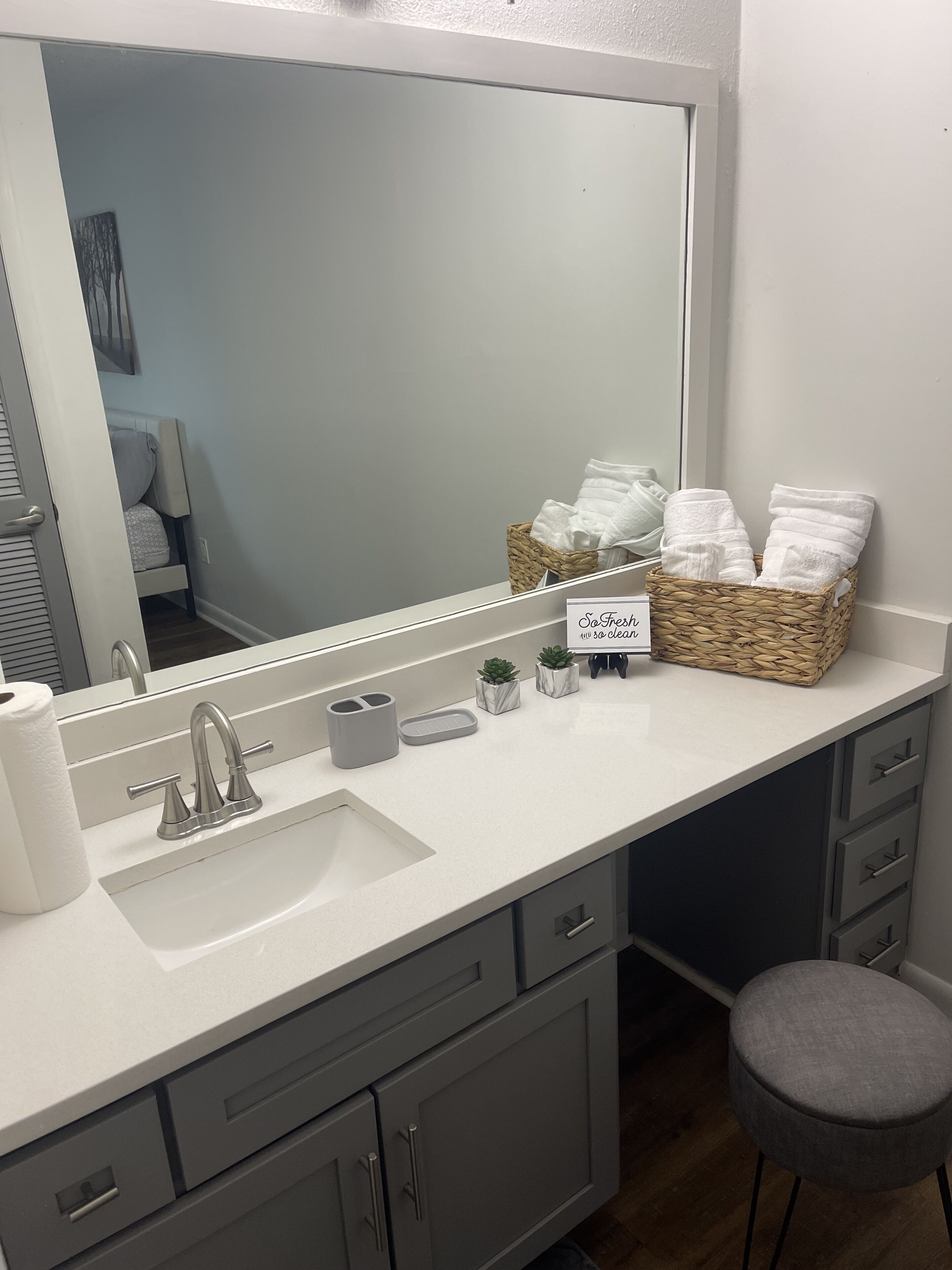 A bathroom with a large mirror, white sink, and grey cabinets. There are two small potted plants on the counter and a basket of towels.