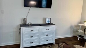 Un salon avec une commode blanche, un téléviseur à écran plat fixé au mur et une petite table sur laquelle est posé un vase.