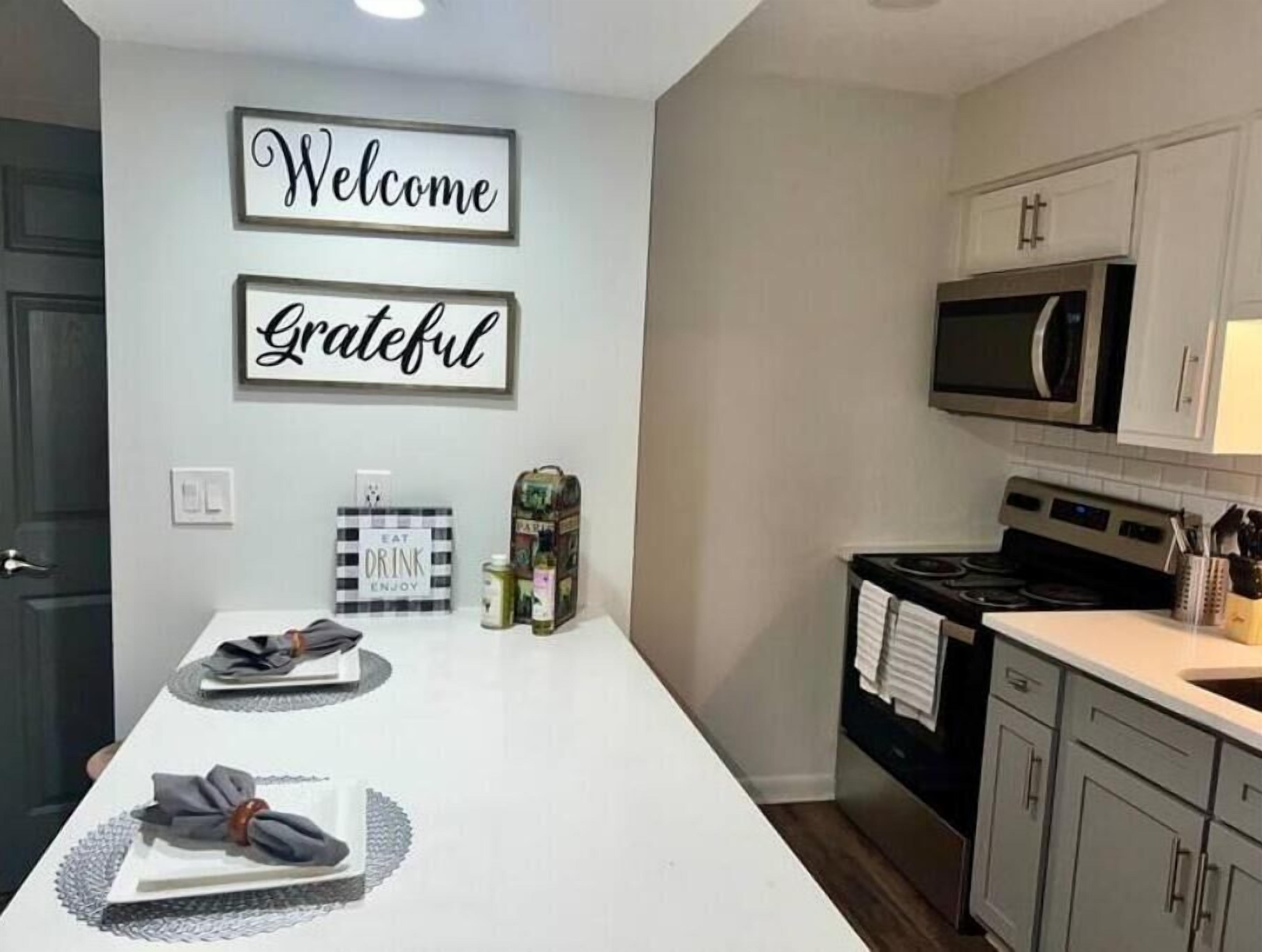 A kitchen with a white countertop, a microwave, cabinets, and a sign that reads "Welcome Grateful".