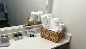 Une salle de bain avec un comptoir blanc, un panier en osier rempli de serviettes blanches, un petit panneau sur lequel on peut lire « Tout frais » et un miroir qui reflète la salle de bain.