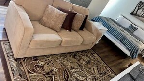 Un canapé beige agrémenté de coussins à motifs, dans un salon où l’on aperçoit un tapis à motifs, un lit avec une literie à rayures et une porte au fond.