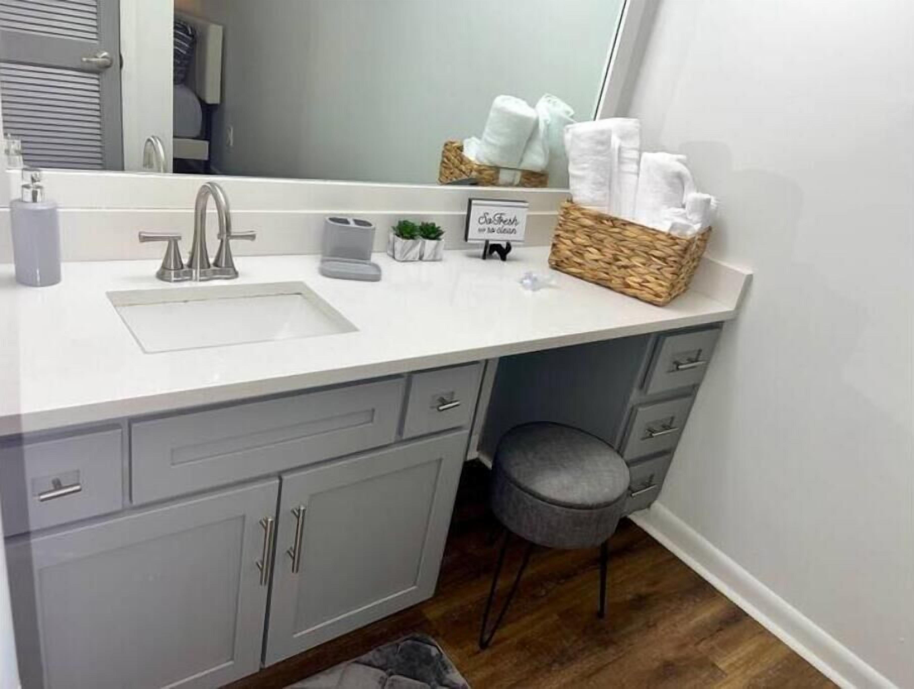 A bathroom with a white sink, grey cabinets, a small stool, and a mirror.