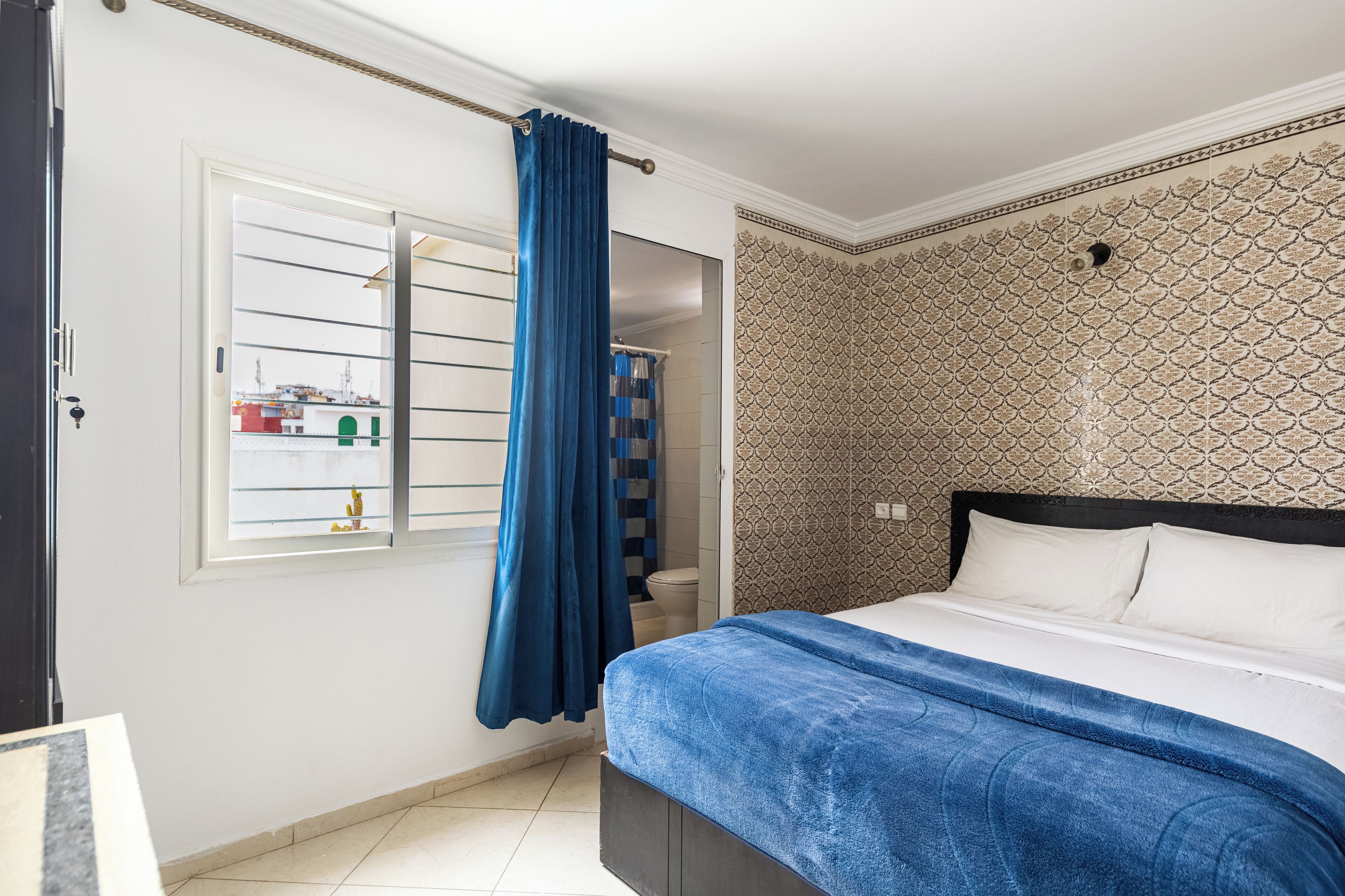 Une chambre à coucher avec un lit, une fenêtre avec des rideaux, un mur à motifs et une salle de bain visible à travers la fenêtre.