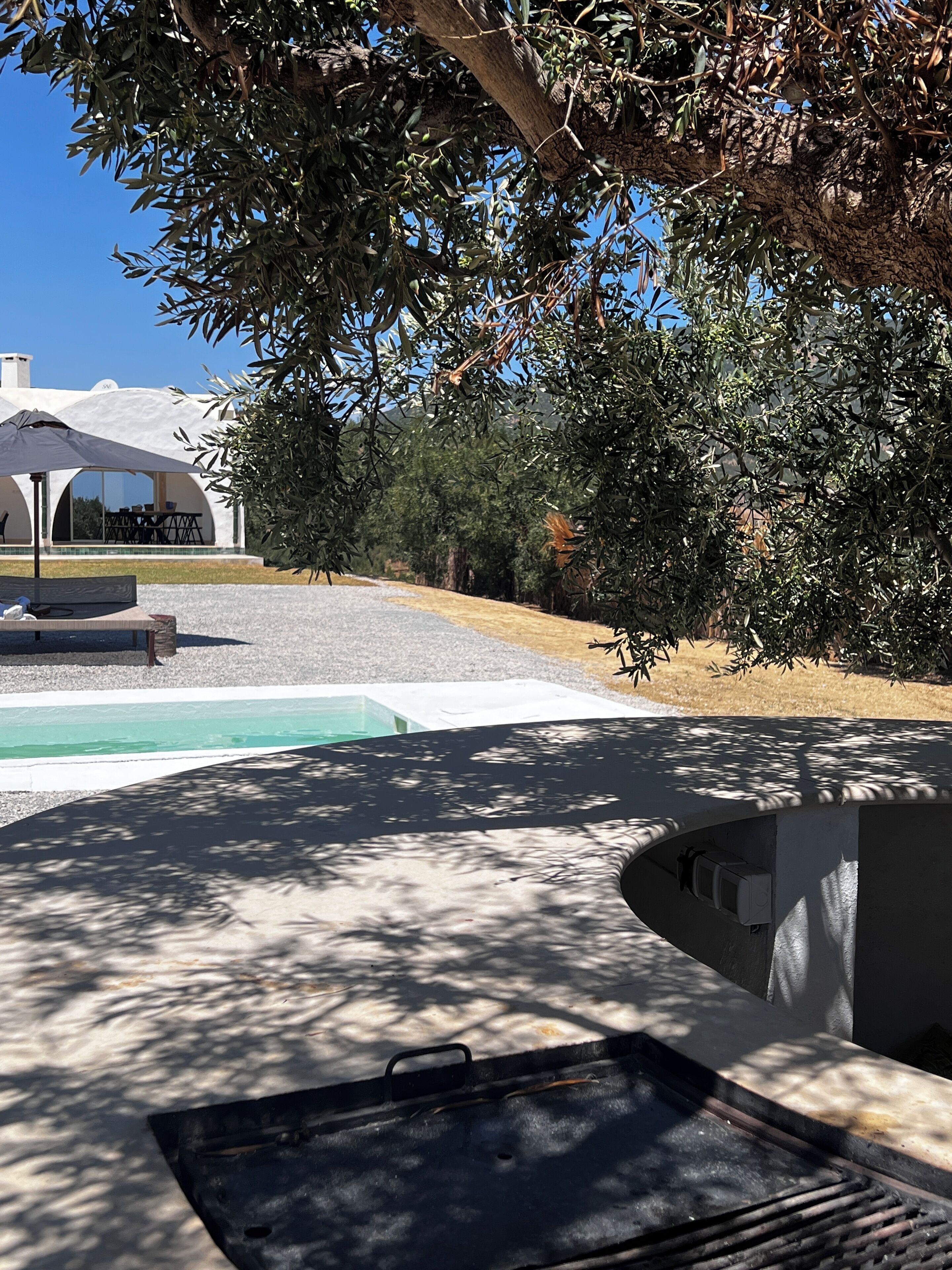 Un espace piscine agrémenté d’une pergola, d’un coin salon couvert et d’un bâtiment à l’extérieur blanc.