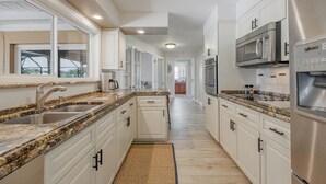 Una cocina moderna con armarios blancos, encimeras de granito, electrodomésticos de acero inoxidable y vistas al exterior.
