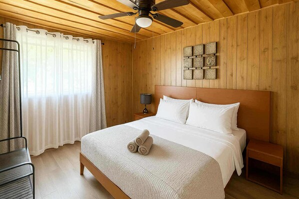 Habitación de hotel con techo de madera, cama con ropa blanca, ventilador de techo y mesita de noche con lámpara.