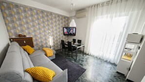 A modern living room with a grey sofa, yellow cushions, a dining table with chairs, a flat-screen TV mounted on the wall, and a kitchen area with a refrigerator and microwave. - Venezia a 15 min bus sotto casa ogni 10 min, parcheggio libero, animali ammessi. (Chirignago-Zelarino)