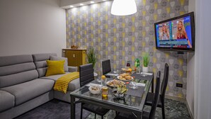 A modern living room with a grey sofa, yellow throw, and a dining table set for two. A TV is mounted on the wall displaying a show. - Venezia a 15 min bus sotto casa ogni 10 min, parcheggio libero, animali ammessi. (Chirignago-Zelarino)