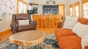 A living room with a brown leather armchair, a wooden coffee table, and an orange sofa. There is a dining area with wooden chairs and a kitchen in the background.