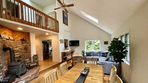 Un vaste espace de vie comprenant une table à manger en bois, une cheminée, une mezzanine dotée d’une rambarde en bois et une grande fenêtre laissant entrer la lumière naturelle.