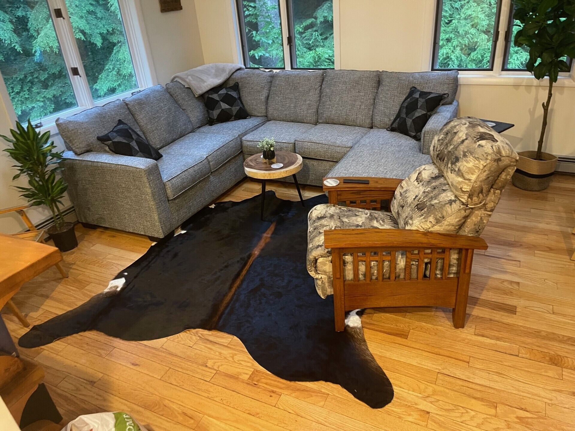 Ein modernes Wohnzimmer mit einer Eckcouch, einem runden Couchtisch und einem gemusterten Sessel. Ein schwarz-weißer Kuhfellteppich liegt auf dem Holzboden.