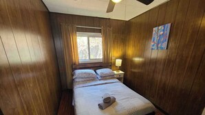 Una habitación con paneles de madera, una cama, una mesita de noche, una lámpara y una ventana con cortinas.
