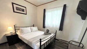 A small, clean room with a single bed, a nightstand with a lamp, a chair, and a window with curtains. - Cottage #7 - 2 Bedroom - Katawoda Cottages (Nobel)
