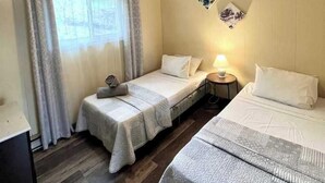 Una habitación de hotel con dos camas individuales, una mesita con una lámpara, una ventana con cortinas y suelo de madera.