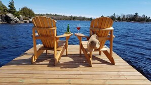 Deux chaises Adirondack en bois, posées sur une jetée en bois, avec un verre de vin rouge et une bouteille contenant un liquide vert.