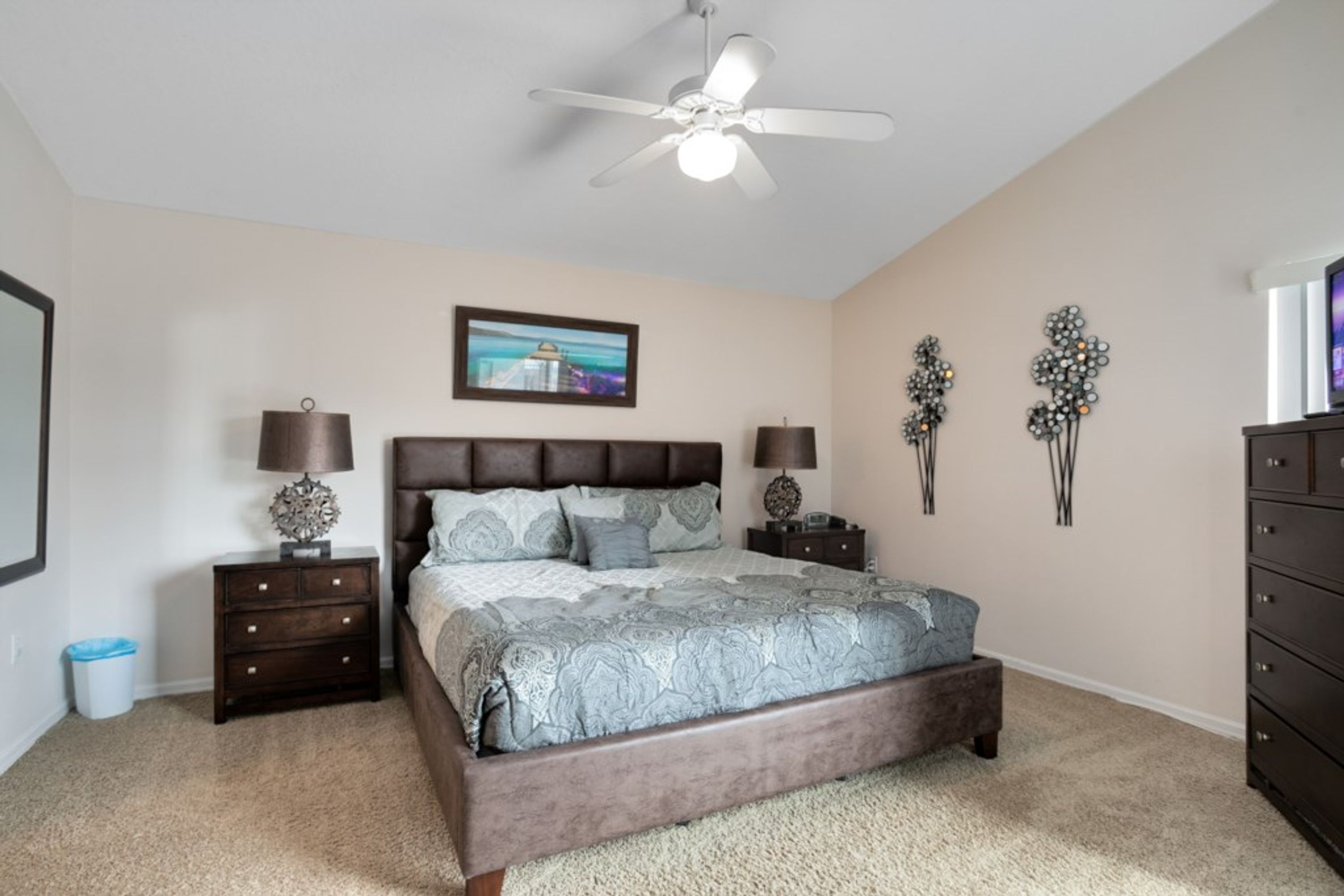 A bedroom with a large bed, two nightstands, a ceiling fan, a wall-mounted TV, and decorative wall art.