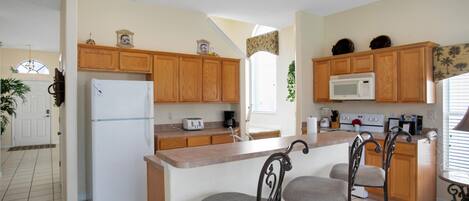 A kitchen with wooden cabinets, a white refrigerator, a microwave, and a bar area with two chairs.