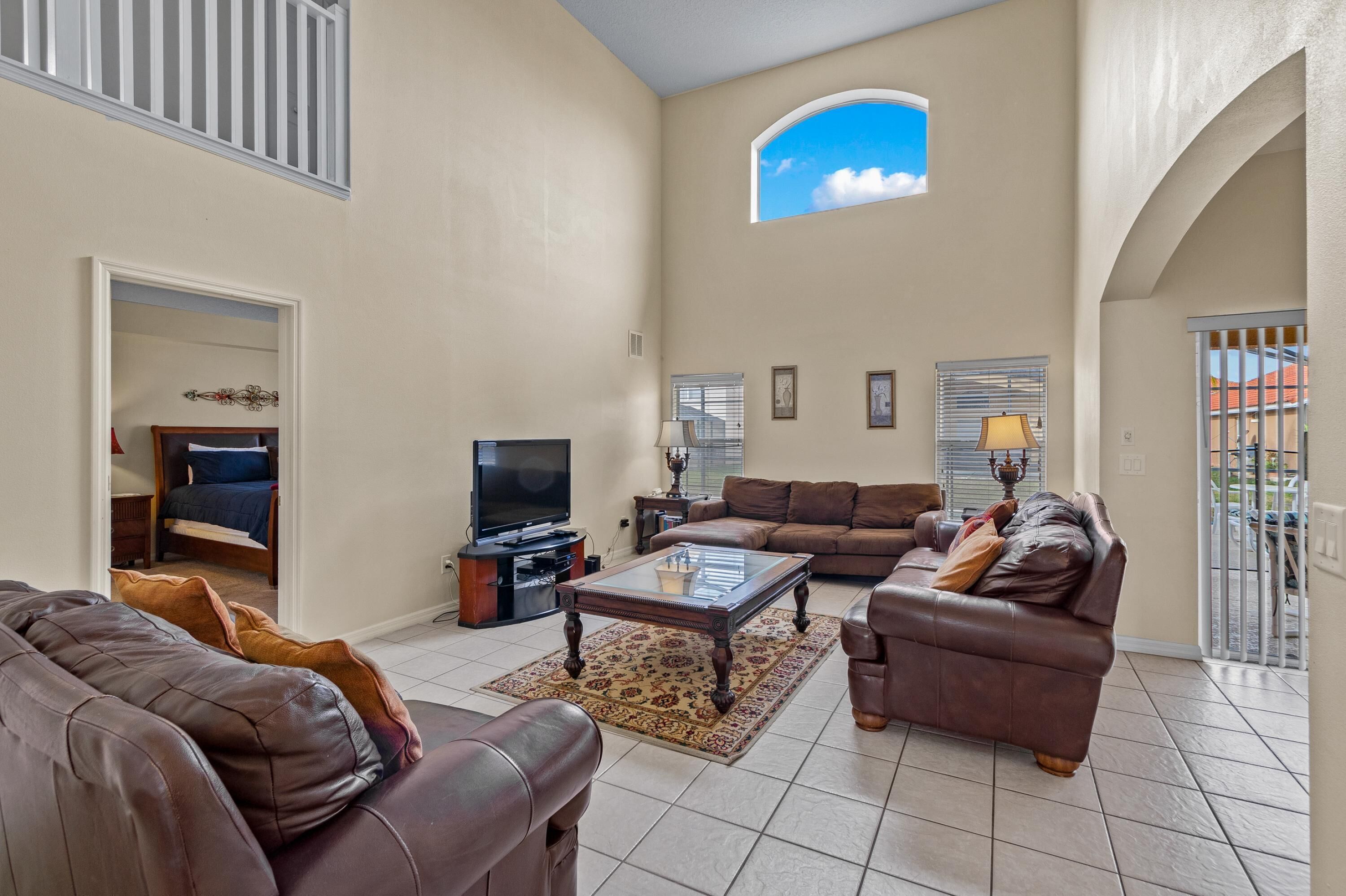 A spacious living room with a large sofa, a coffee table, and a television. There is a balcony with a view of the sky and a gate.