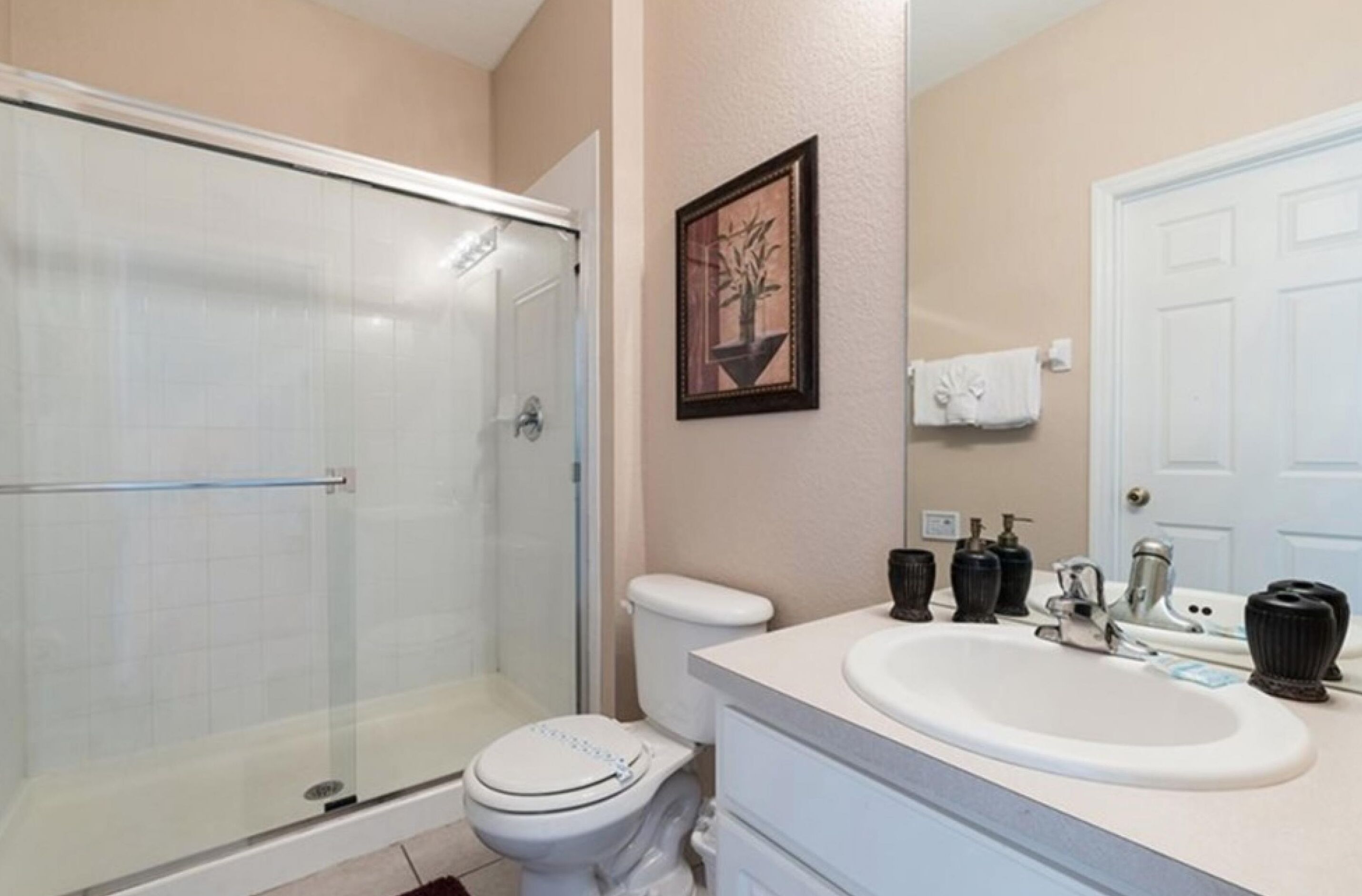 A bathroom with a glass-enclosed shower, a toilet, a sink with a mirror, and a framed picture on the wall.