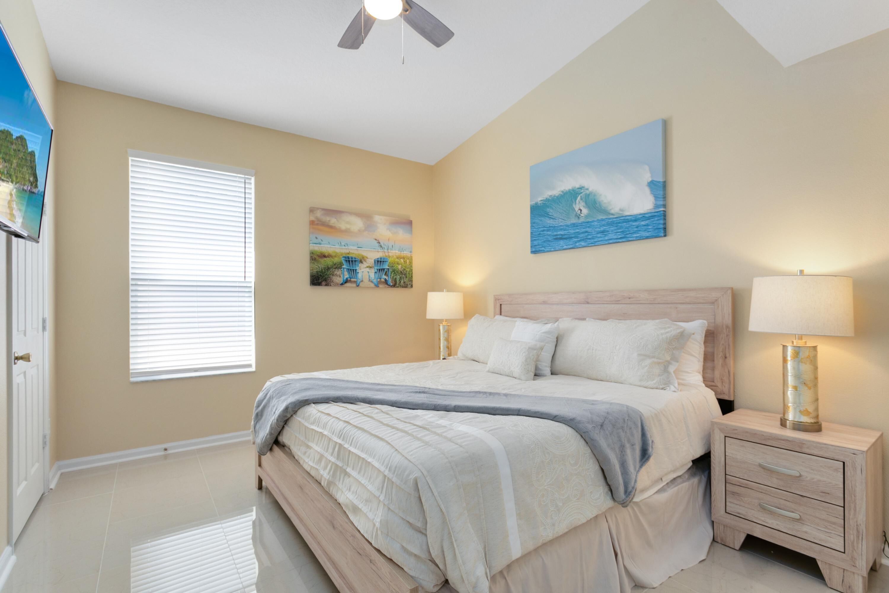 A bedroom with a bed, two bedside tables, a ceiling fan, a window with blinds, and wall art.