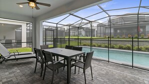 Une terrasse extérieure avec une piscine, protégée par une structure en verre, comprenant une table, des chaises et un ventilateur de plafond.