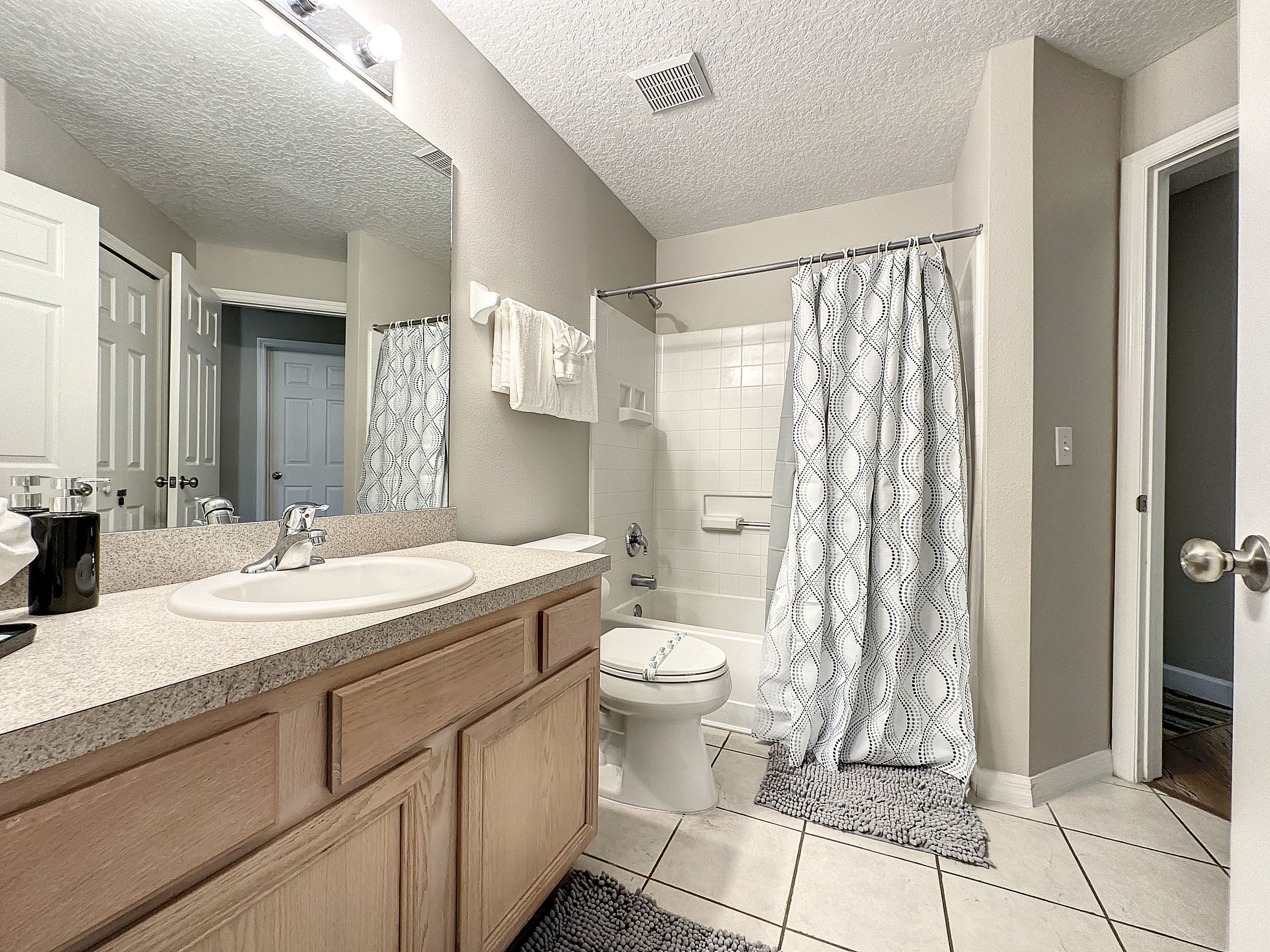 A bathroom with a double sink vanity, a toilet, a shower with a patterned curtain, and a door leading to another room.