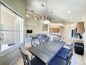 A spacious living area with a large dining table, comfortable seating, a kitchen, and a balcony with a sliding door.