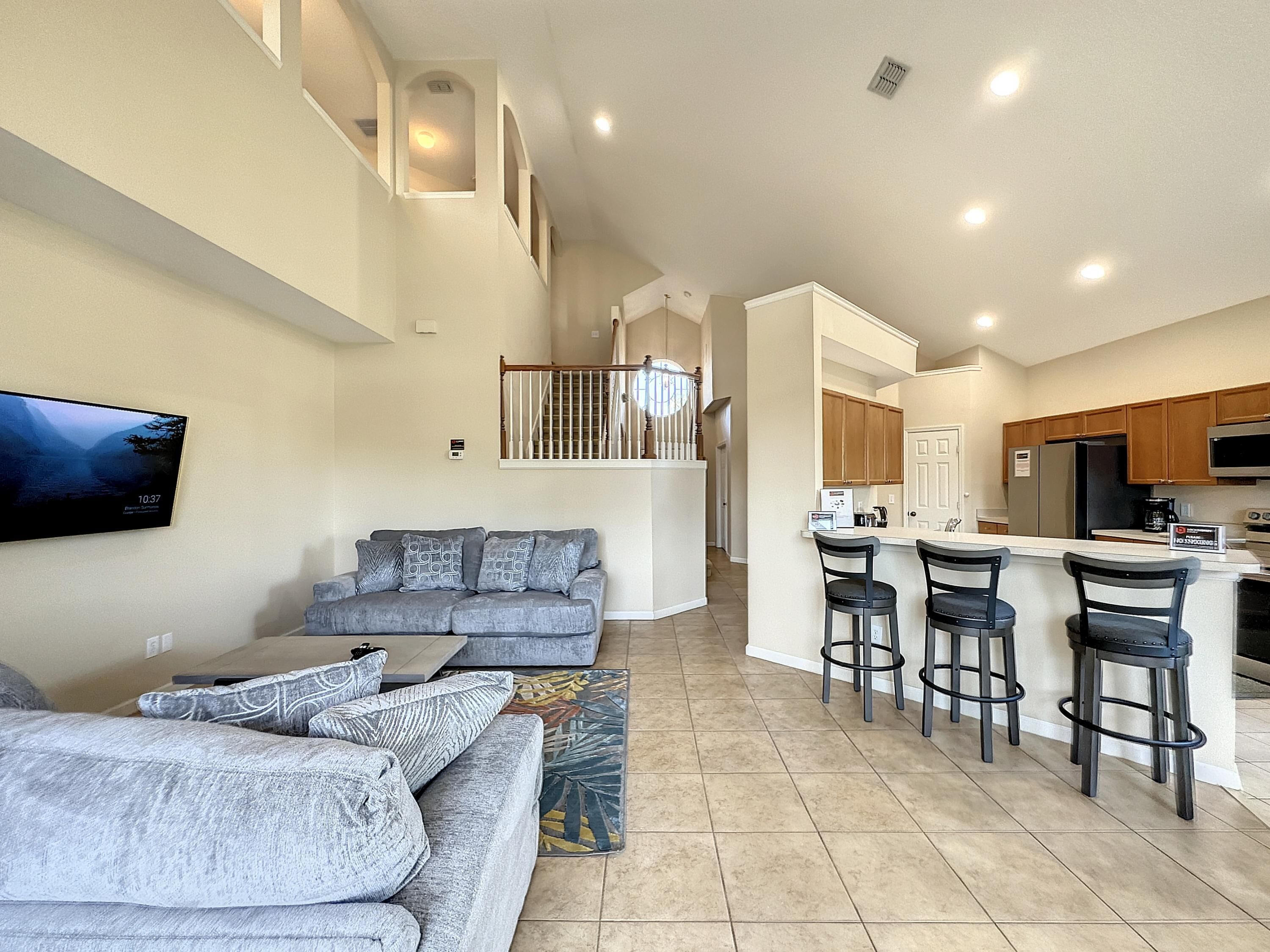 A spacious living area with a grey sofa, a flat-screen TV mounted on the wall, and a kitchen area with wooden cabinets and a stainless steel refrigerator.