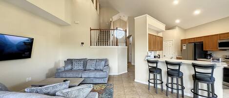 A spacious living area with a grey sofa, a flat-screen TV mounted on the wall, and a kitchen area with wooden cabinets and a stainless steel refrigerator.