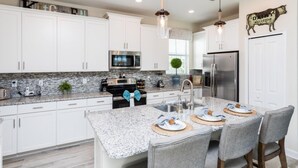 A modern kitchen with white cabinets, granite countertops, stainless steel appliances, and a dining area with bar stools. - 5 Bedroom Home is Ideal for an Elegant Vacation (Kissimmee)