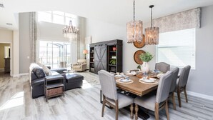 A spacious living room with a dining area, featuring a large wooden table, grey upholstered chairs, and a chandelier. - 5 Bedroom Home is Ideal for an Elegant Vacation (Kissimmee)