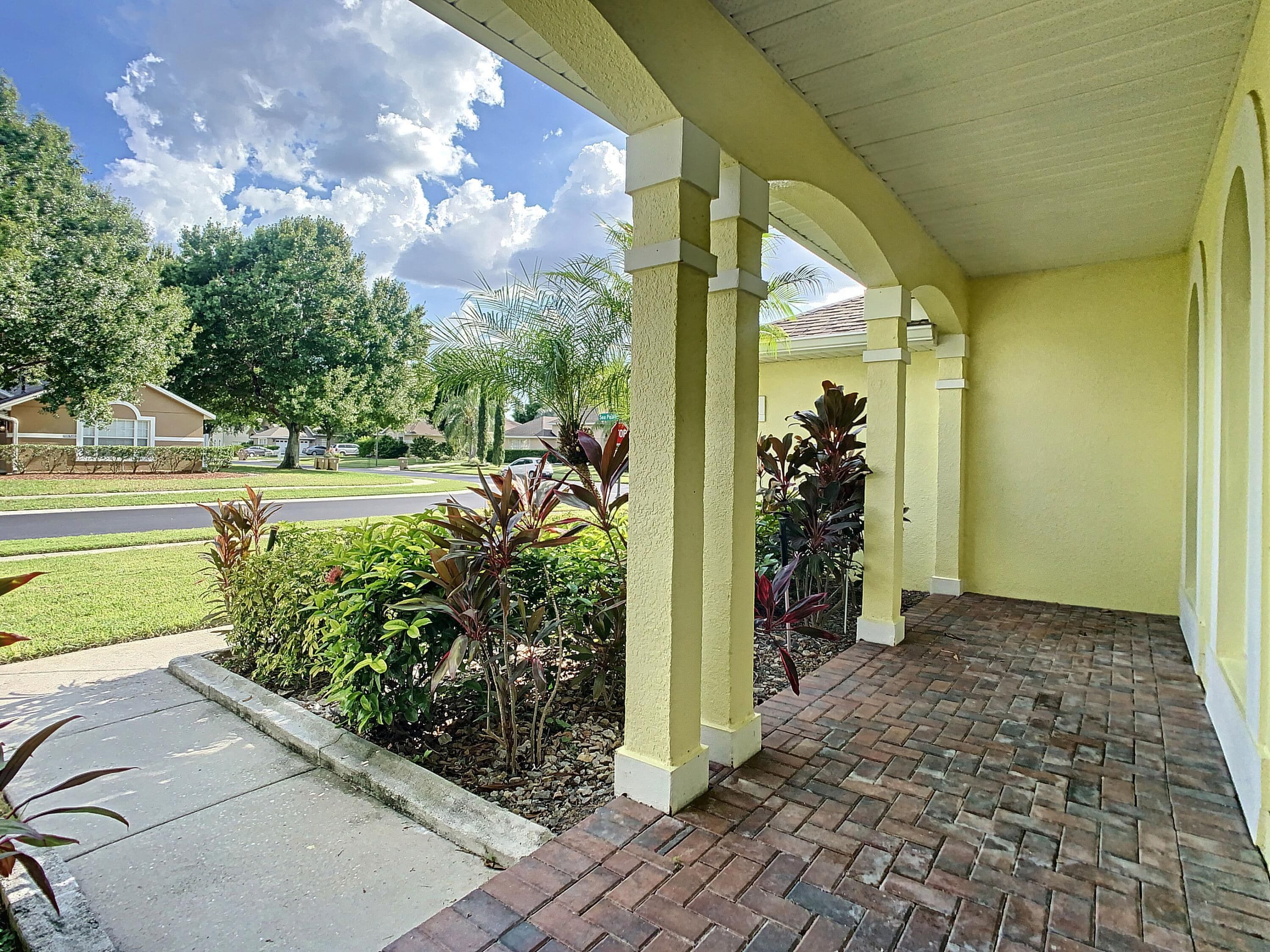 A covered patio area with brick flooring and yellow walls, featuring a view of a residential street and greenery.