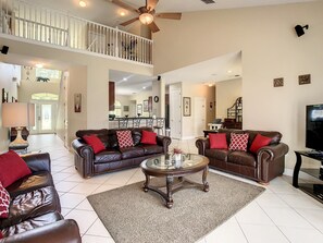A spacious living room with a high ceiling, a ceiling fan, a dining area in the background, and a staircase leading to an upper level. - Fantastic 7 Bed 6 Bath in Prestigious Gated Comm (Kissimmee)