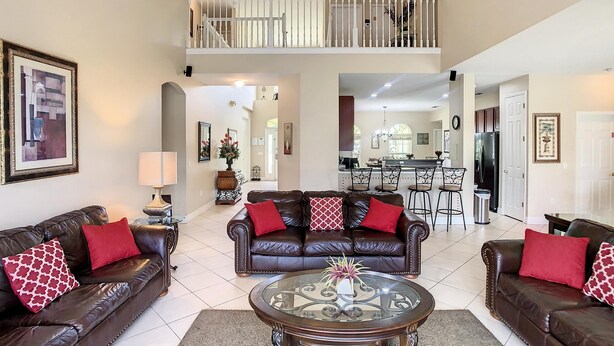 A spacious living room with two leather sofas, a glass coffee table, and a ceiling fan. A kitchen and dining area are visible in the background. - Fantastic 7 Bed 6 Bath in Prestigious Gated Comm (Kissimmee)